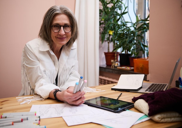 Portrait of a mature pretty woman fashion designer at workplace with clothes sketches laptop color wheels on a digital tablet in a tailoring atelier