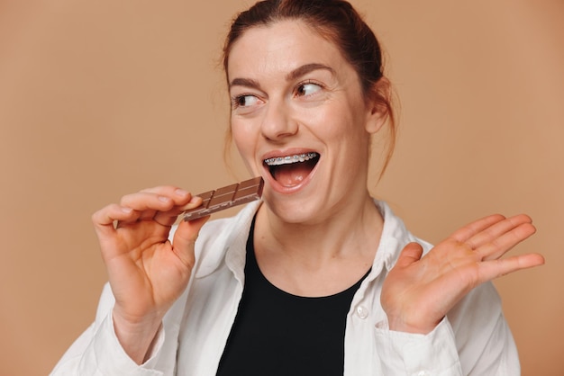 Portrait of mature woman in braces biting chocolate on a beige background