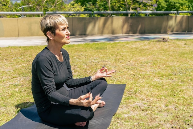 Портрет зрелой женщины, сидящей в позе лотоса, практикующей йогу в общественном парке в солнечный день. Концепция здорового образа жизни