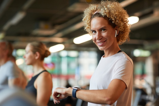 Portrait of mature woman with curly hair smiling at camera while checking her pulse on wristwatch during training