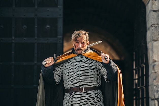 Портрет средневекового старшего воина в доспехах после битвы с кровью на лице. Воин держит в руках два меча