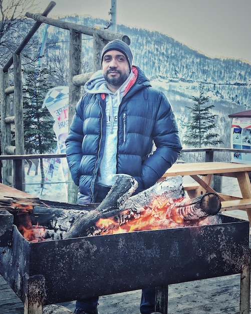 Foto ritratto di un uomo adulto con le mani in tasca in piedi vicino a una fossa di fuoco contro la montagna durante l'inverno