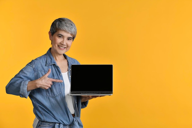 Портрет азиатской женщины средних лет 50-х годов в повседневной джинсовой рубашке, белой футболке, держащей ноутбук и указывающей пальцами на желтом фоне, смотрящей в камеру