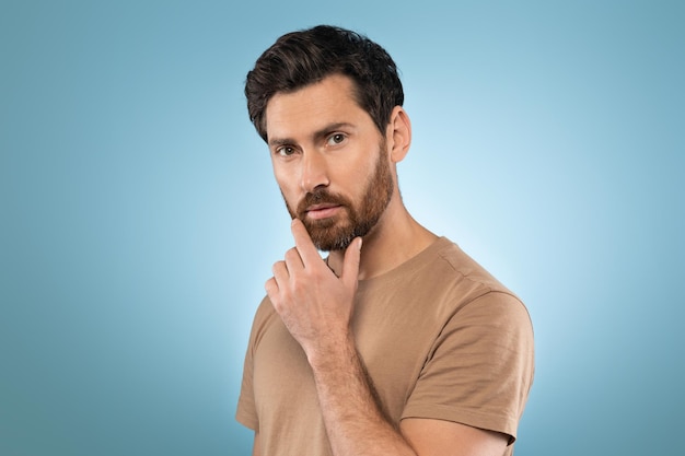 Portrait of middle aged bearded man posing and touching chin standing over blue studio background