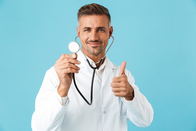 Portrait of middle-aged medical specialist wearing white coat holding stethoscope, standing isolated over blue wall