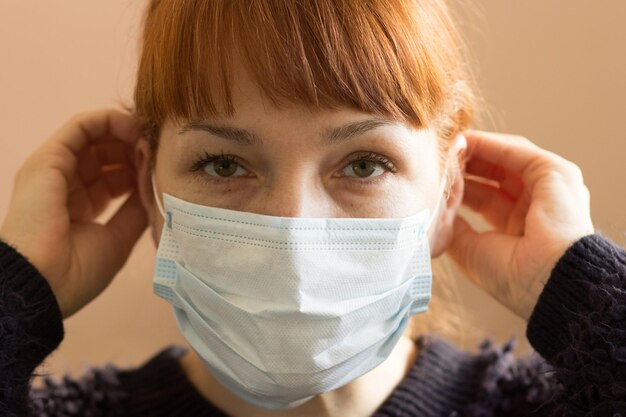 Portrait of middle aged woman putting on surgical mask on face and looking at camera indoor