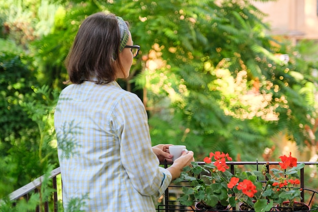 Ritratto di donna di mezza età che si rilassa su un balcone paesaggistico aperto con una tazza di caffè, donna che si gode sulla terrazza con piante, spazio di copia