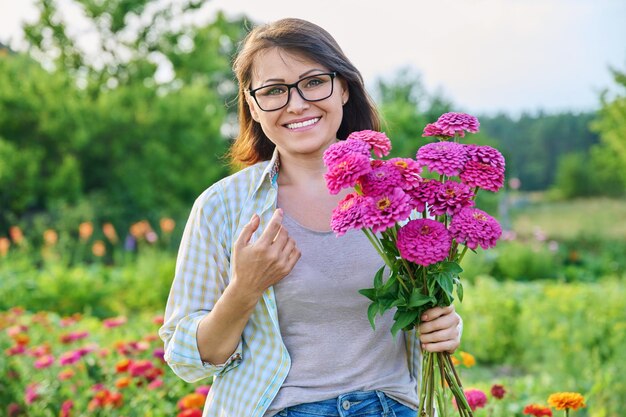 屋外の花の花束を持つ中年女性の肖像画、夏の日に百日草の花を持ったカメラを見て笑顔の幸せな 40 代の女性自然美成熟人のコンセプト