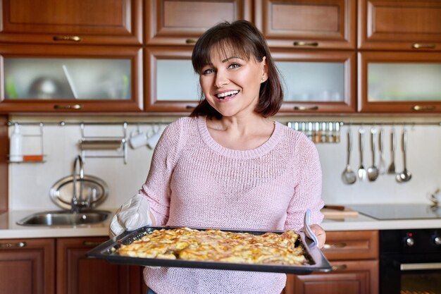 Portrait of middle aged woman with pan of cooked oven baked meat looking at camera at home in kitchen Cooking at home recipe meat under cheese people concept