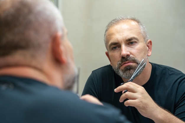 Portrait of middleaged handsome man cutting his beard with scissors selfcare during quarantine