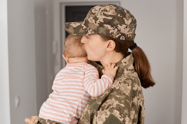 Portrait of military mother wearing camouflage uniform and hat,\
moms arrival after served in army, female kissing her baby, missing\
so much while being absent at home.