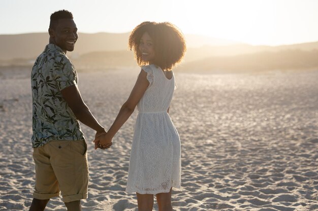 Портрет пары смешанной расы, которая вместе проводит свободное время на пляже в солнечный день, держась за руки и улыбаясь в камеру, а за ними сияет солнце.