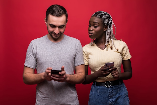 Ritratto di uomo di razza mista e donna accigliata e sbirciare a vicenda i telefoni cellulari isolati su sfondo rosso