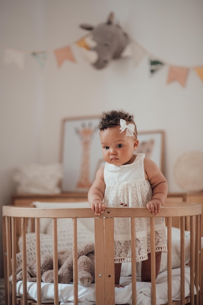 Portrait of mixed race toddler girl in round wooden baby crib at cozy childrens room neutral tones