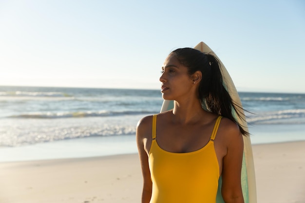 海に向かってビーチでサーフボードを持つ混血女性のポートレート。海辺での健康的なアウトドアレジャータイム。