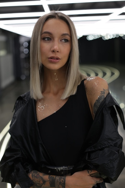 Portrait of a model girl in a black bodysuit windbreaker and bright silver sequins on the body which stands indoors with bright artificial lighting on the ceiling and looks to the side