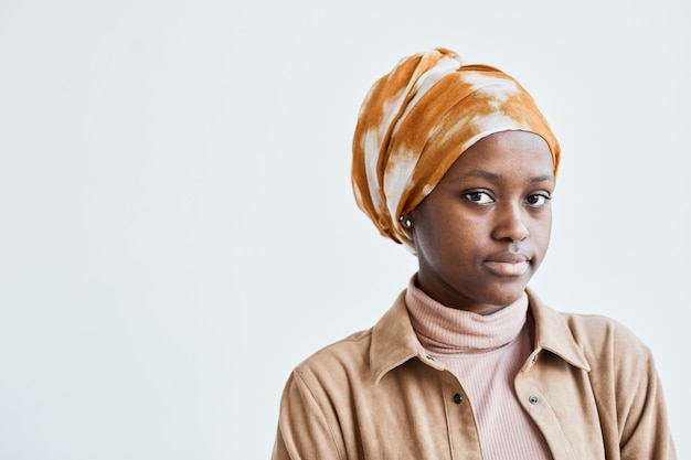 Portrait of modern African-American woman looking at camera and wearing headscarf while posing against white wall, copy space