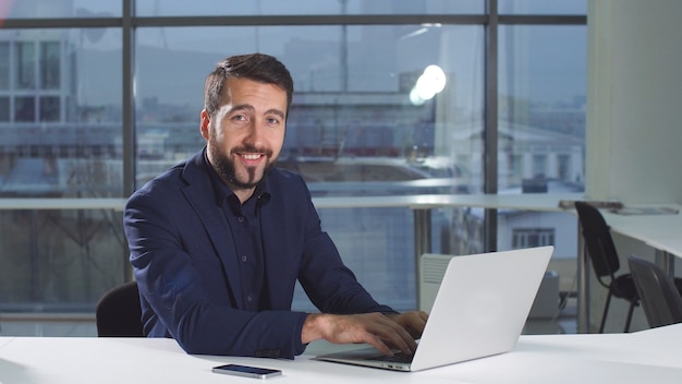 Portrait of modern attractive businessman working at desk in office using laptop