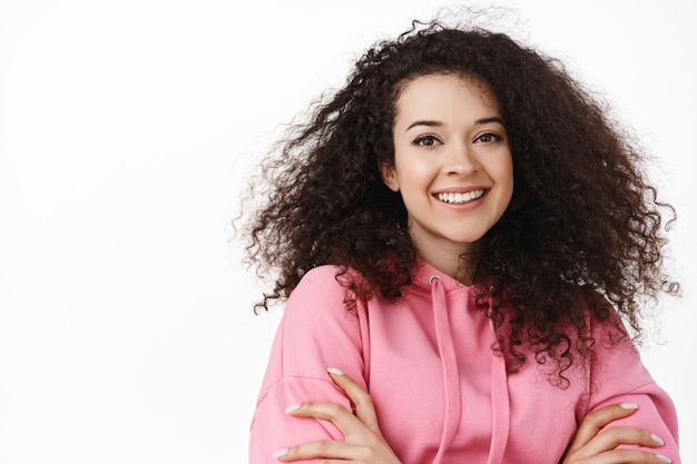 Portrait of of modern brunette girl with natural curly hair, cross arms on chest, smiling white teeth, looking happy and relaxed, standing on white