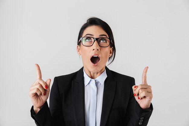 Portrait of modern businesswoman 30s in formal wear and eyeglasses pointing fingers upward, isolated over white wall