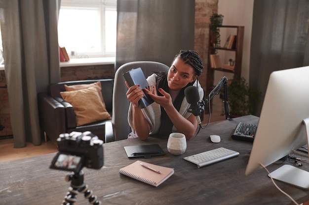 Portrait of modern female influencer wearing sitting at desk in front of camera demonstrating book a