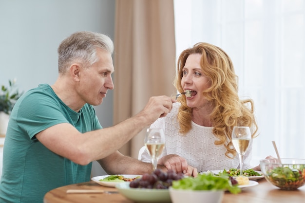 Portrait of modern mature couple enjoying dinner together at home, man giving wife piece of food on fork, copy space