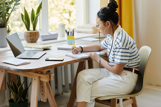 Portrait of modern teenage girl studying at desk at home and writing in notebook, copy space