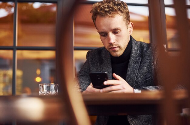 Portrait of modern young guy in formal clothes that sits in the cafe with phone in hands.