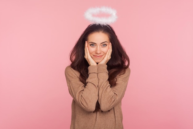 Portrait of modest young pretty woman with halo above head holding hands on face and looking at camera with kind timid smile angelic beauty shyness indoor studio shot isolated on pink background