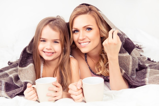 Portrait of mom and daughter basking under plaid with cups of hot milk