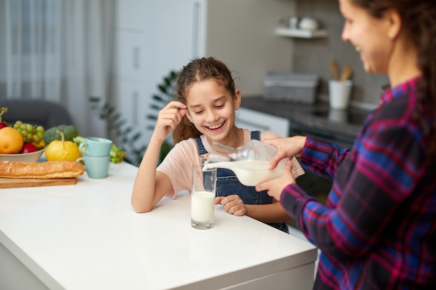 Portrait of a mom pours her cute laughing daughter milk in glass for breakfast