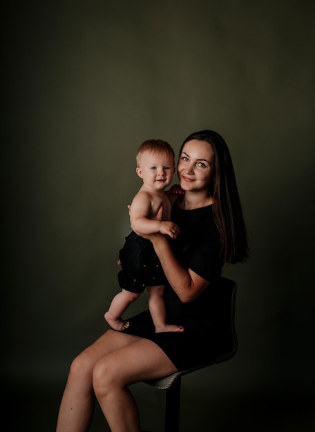 Portrait of a mother in a black dress sitting on a chair with her baby daughter on a green isolated background with a place for text