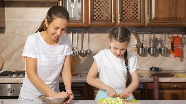 Portrait of mother and daughter are cooking apple pie together in the kitchen, mom is kneading a dough and her teen daughter is cutting apple.