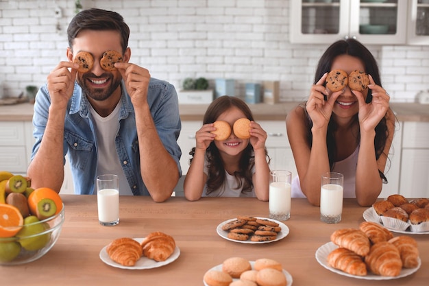 Photo portrait of mother daughter and father covering eyes with cookies on the kitchen