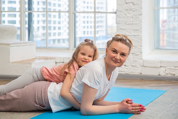 portrait of mother and daughter in sportswear lying on a yoga mat in front of  window, hugging 