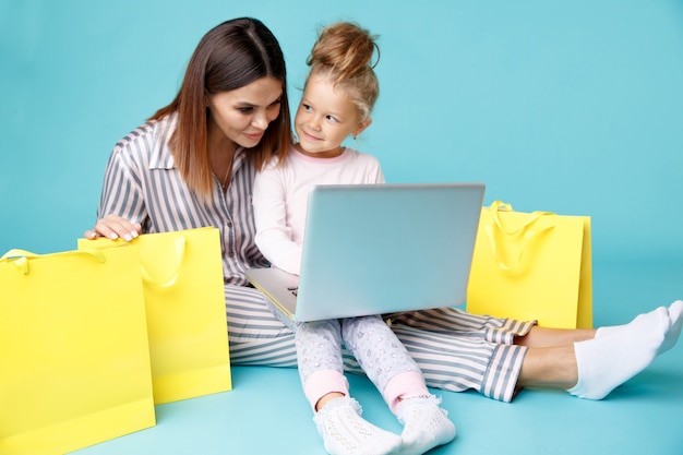 Portrait of mother and daughter with laptop sitting on the floor isolated over the blue studio. online family shopping concept.