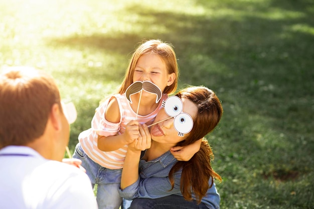 Ritratto di madre padre e figlia che si divertono insieme nel parco usando maschere di carta sui bastoncini