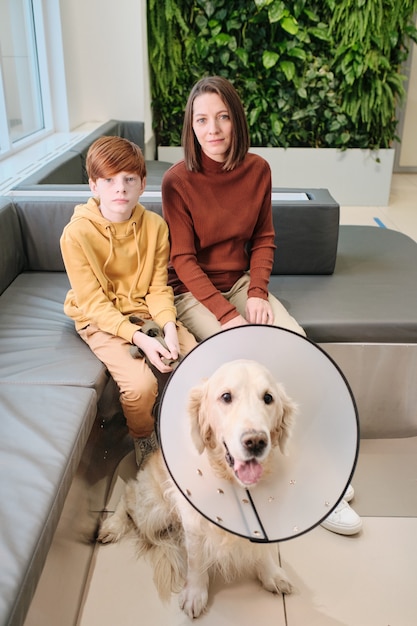 犬と一緒にソファに座って、医者の訪問を待っているカメラを見ている母と彼女の息子の肖像画
