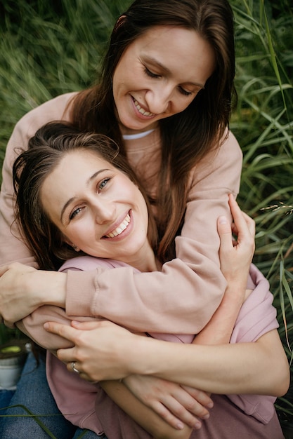 Portrait of mother hugging beautiful adult daughter, sitting on green grass, dressed in beige sweaters