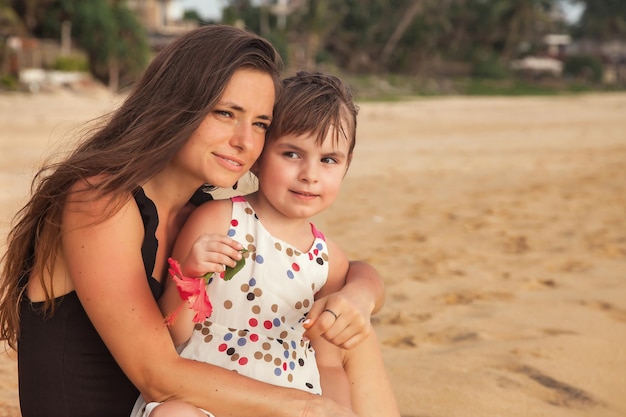 Madre del ritratto e piccola figlia insieme che si siedono sulla spiaggia sabbiosa tropicale al fondo delle palme