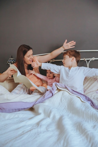 Portrait of mother reading story book to her daughter and son kids sitting in the bed. Weekend family leisure time concept.