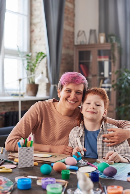 Portrait of mother and son smiling at camera while painting eggs at the table and celebrating Easter at home