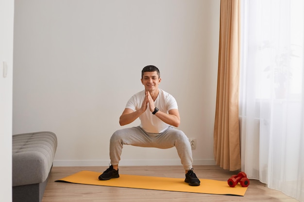 Portrait of motivated athletic man wearing sportswear doing squat, lower body sport exercise, warming up and training muscles for strength and flexibility, keeps palms together, looking at camera.