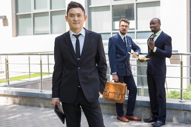 Photo portrait of multi ethnic business team. three happy smiling men standing against the backdrop of the city.  the foreground of a chinese  man  with laptop