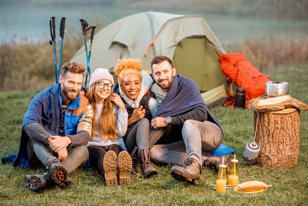 Portrait of multi ethnic group of friends dressed in sweaters hugging together and warming up in plaid at the camping during the evening