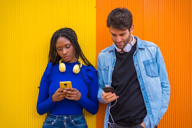 黄色の背景の大学キャンパスで携帯電話を見ている白人男性と黒人女性の多民族カップルのポートレート
