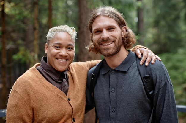 Photo portrait of multiethnic happy couple embracing and smiling at camera while walking in the forest