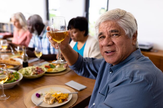 Photo portrait of multiracial senior man enjoying wine while having lunch with friends at dining table. happy, alcohol, meal, nursing home, unaltered, togetherness, support, assisted living, retirement.