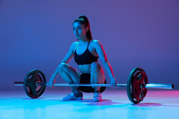 Portrait of muscled woman in sportswear training with a barbell isolated on purple background in neon light