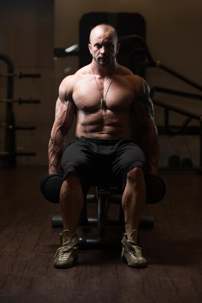 Портрет мускулистого мужчины, отдыхающего на скамейке в тренажерном зале с гантелями